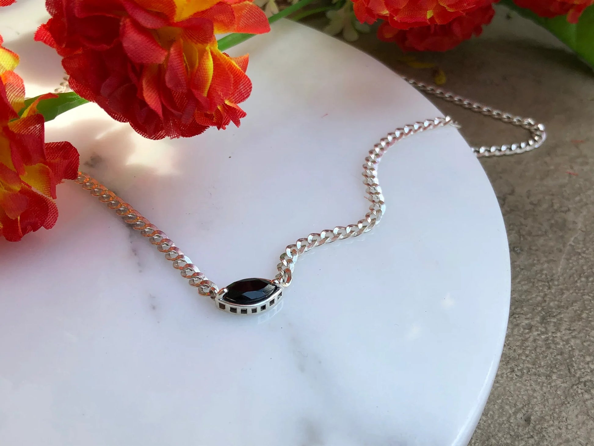 Silver necklace with a black pendant on a marble surface with red flowers in the background