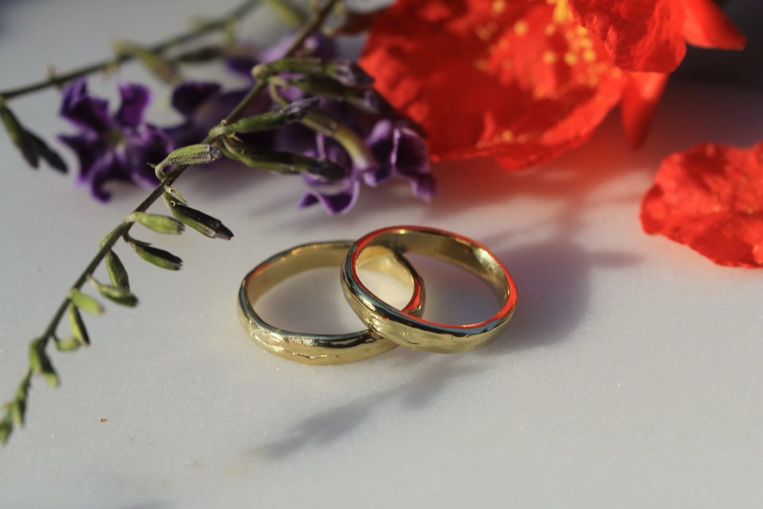 Two gold rings on a white surface with purple and red flowers in the background.